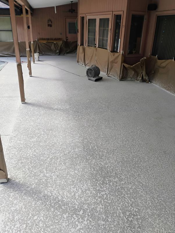 Covered patio with fresh gray flooring, wooden posts, windows, and a floor fan in the background.