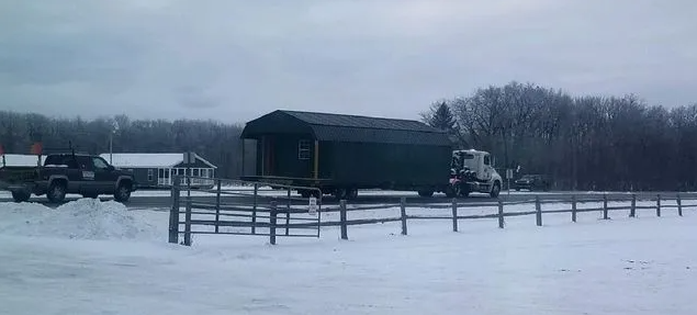 Truck transporting a dark green shed on a snowy day near a fence.