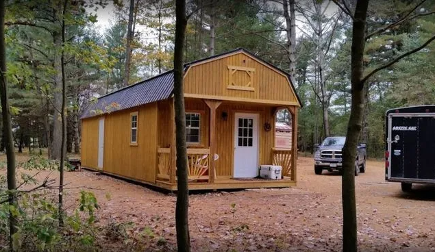 Yellow cabin with porch in a wooded area, parked vehicles nearby.