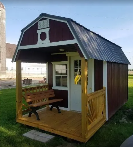 Small maroon and white shed with porch, bench, and metal roof on a grassy field.