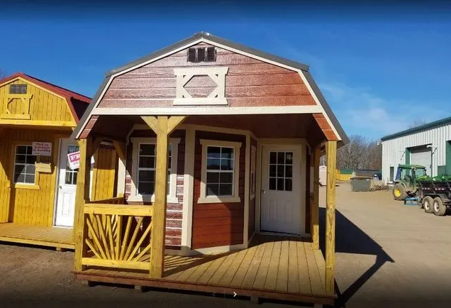 Red and tan wooden shed with porch, white door and window, displayed outdoors on a sunny day.