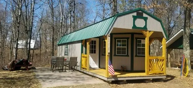 A small, light-colored cabin with a green roof and yellow porch in a wooded area; an American flag hangs on the porch.