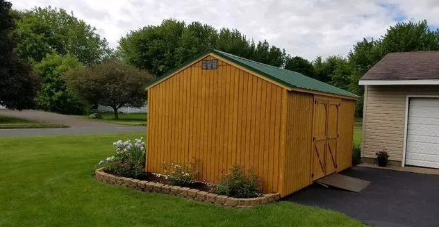 Yellow shed with green roof, beside a driveway and garden, set in a grassy yard, cloudy sky.