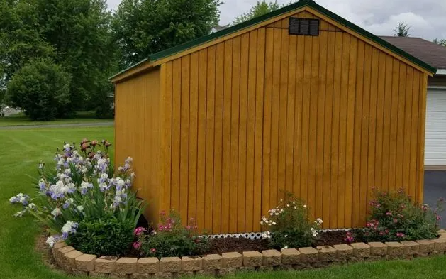 Yellow wooden shed with a green roof, surrounded by a flower bed.