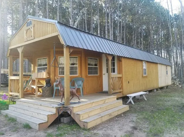Wooden cabin with porch and steps, surrounded by trees in a grassy area.