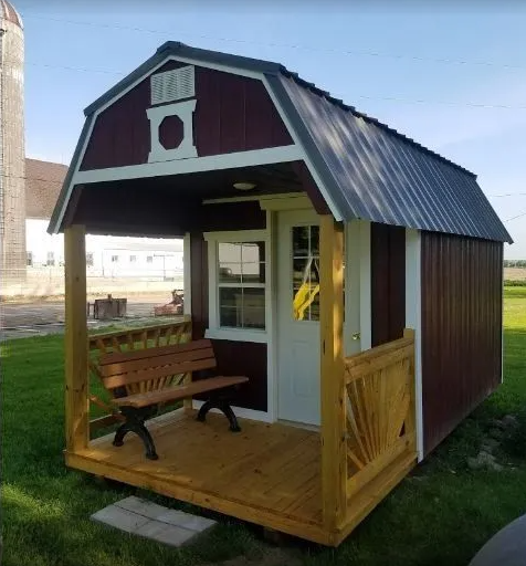 Red and white shed with porch, wooden bench, and metal roof, on a grassy field.