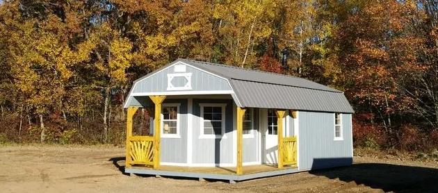 A small, gray shed with a porch and yellow accents, surrounded by autumn trees.