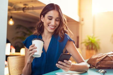 Woman smiling at a café table while holding a coffee cup and looking at her phone.