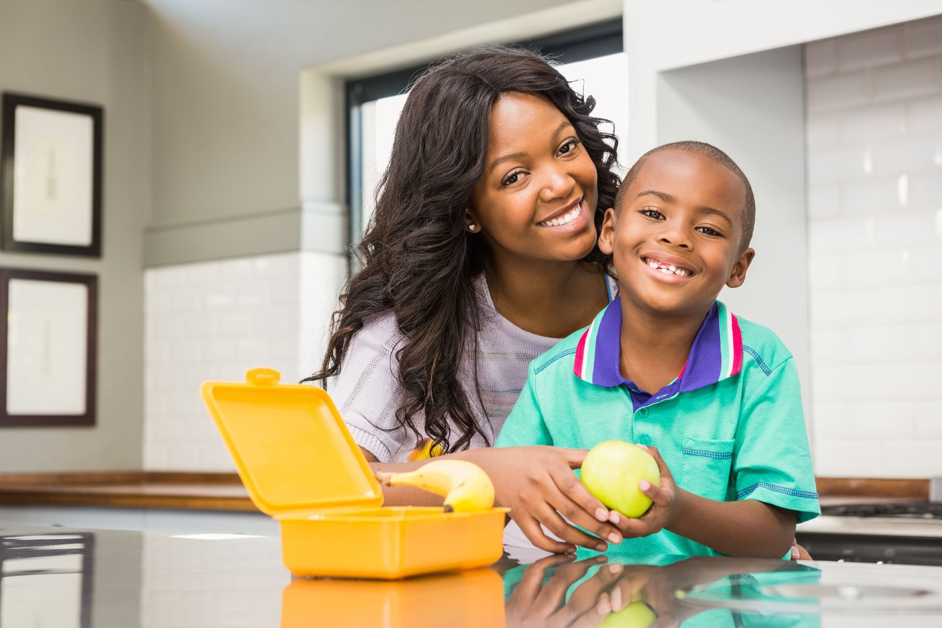 Woman and child smiling in a kitchen, with a yellow lunchbox and a green apple on the counter.