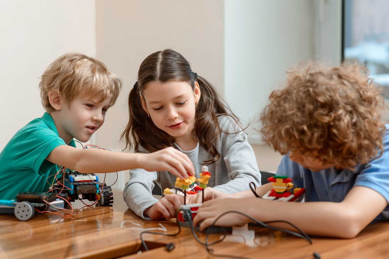 Three children playing with toy cars and wires on a wooden table indoors