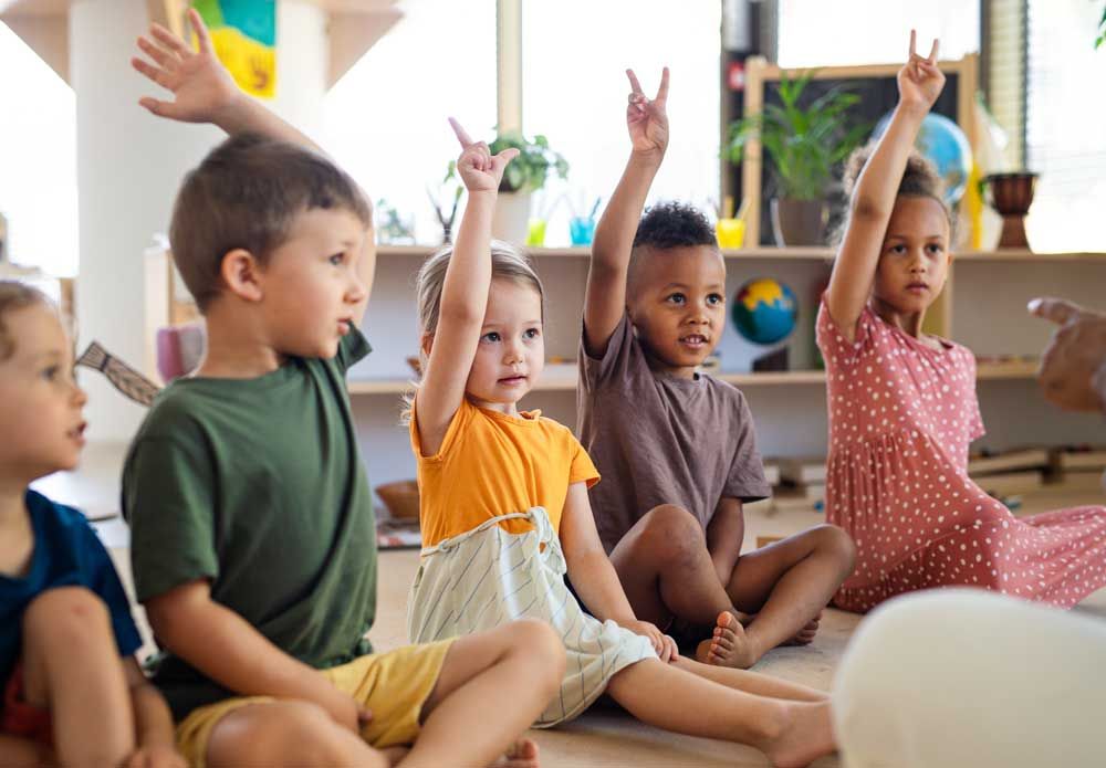 Children sitting cross-legged in a classroom with hands raised during an activity
