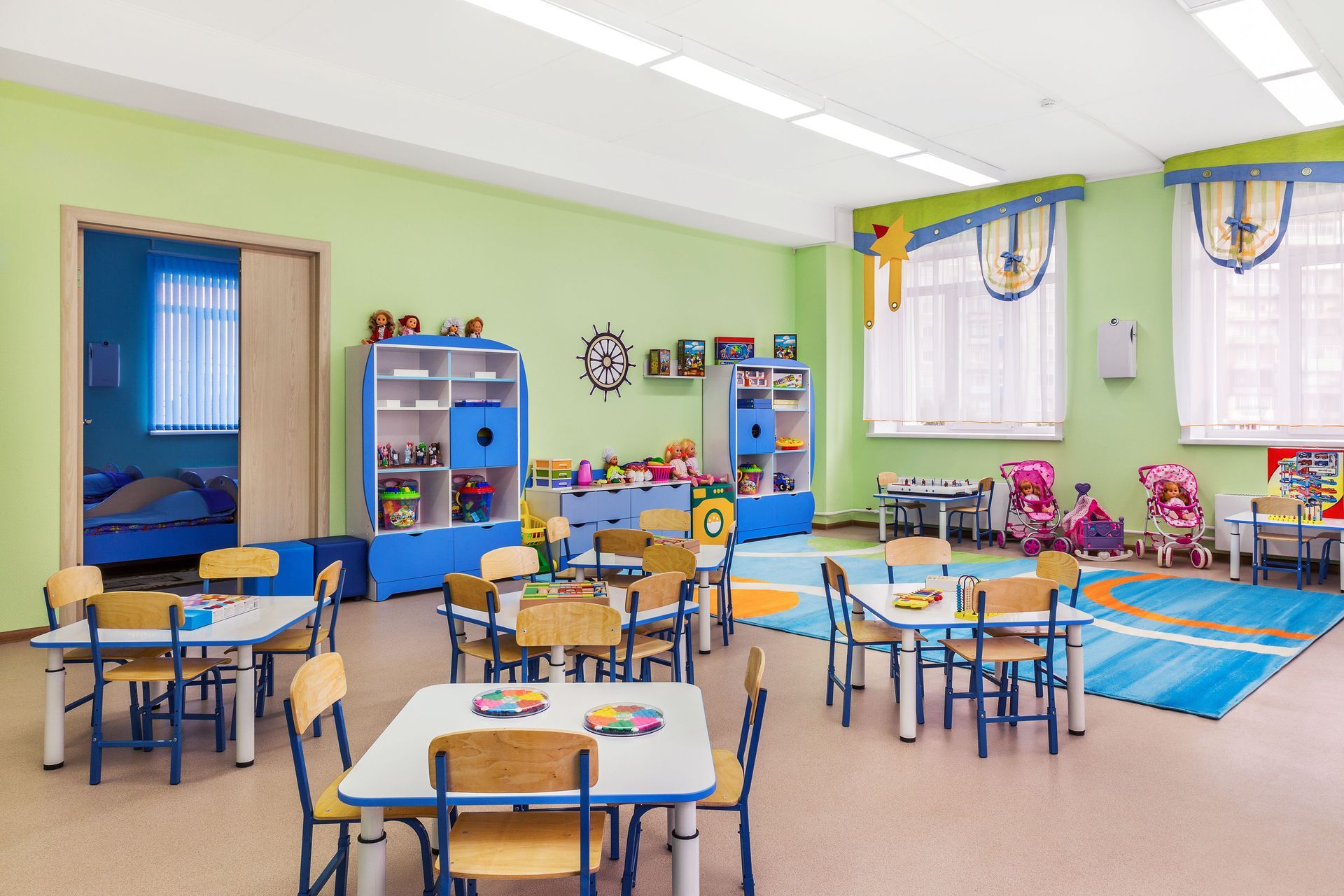 Bright classroom with blue tables and chairs, colorful wall decor, and windows with curtains.