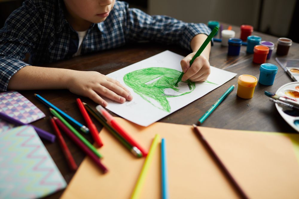 Child drawing a green leaf with crayons and paint jars on a table