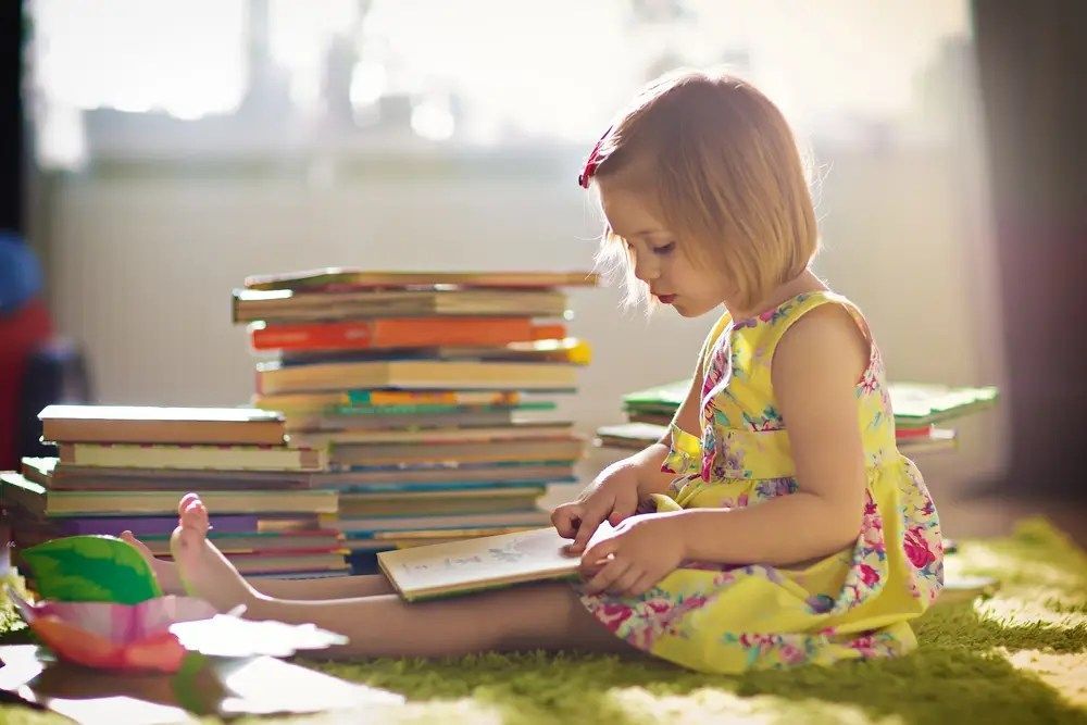Child reading on grass beside colorful stacks of books in bright sunlight