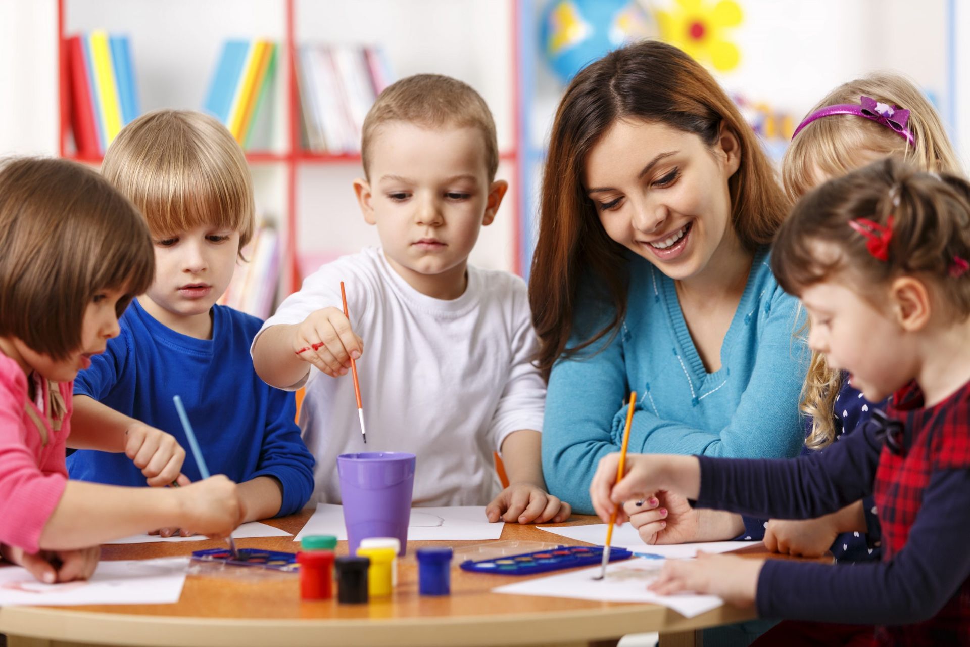 Teacher and children painting at a classroom table with colorful art supplies