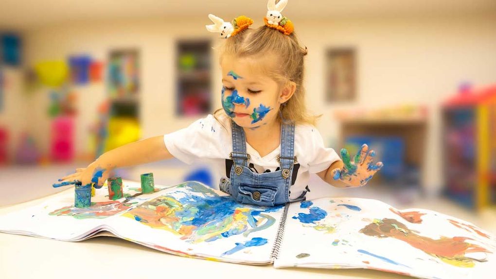 Young child painting at a table with colorful paint splashes in a bright classroom