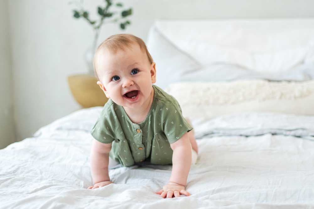 Smiling baby on a white bed, wearing a green outfit and crawling toward the camera.