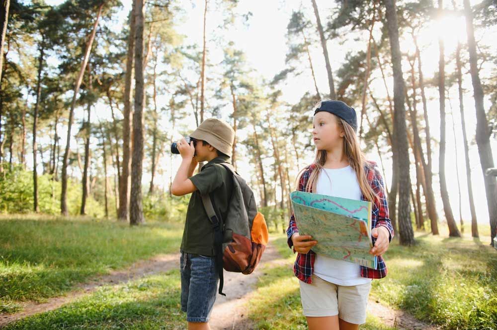 Two children with backpacks and maps hiking on a sunlit forest trail