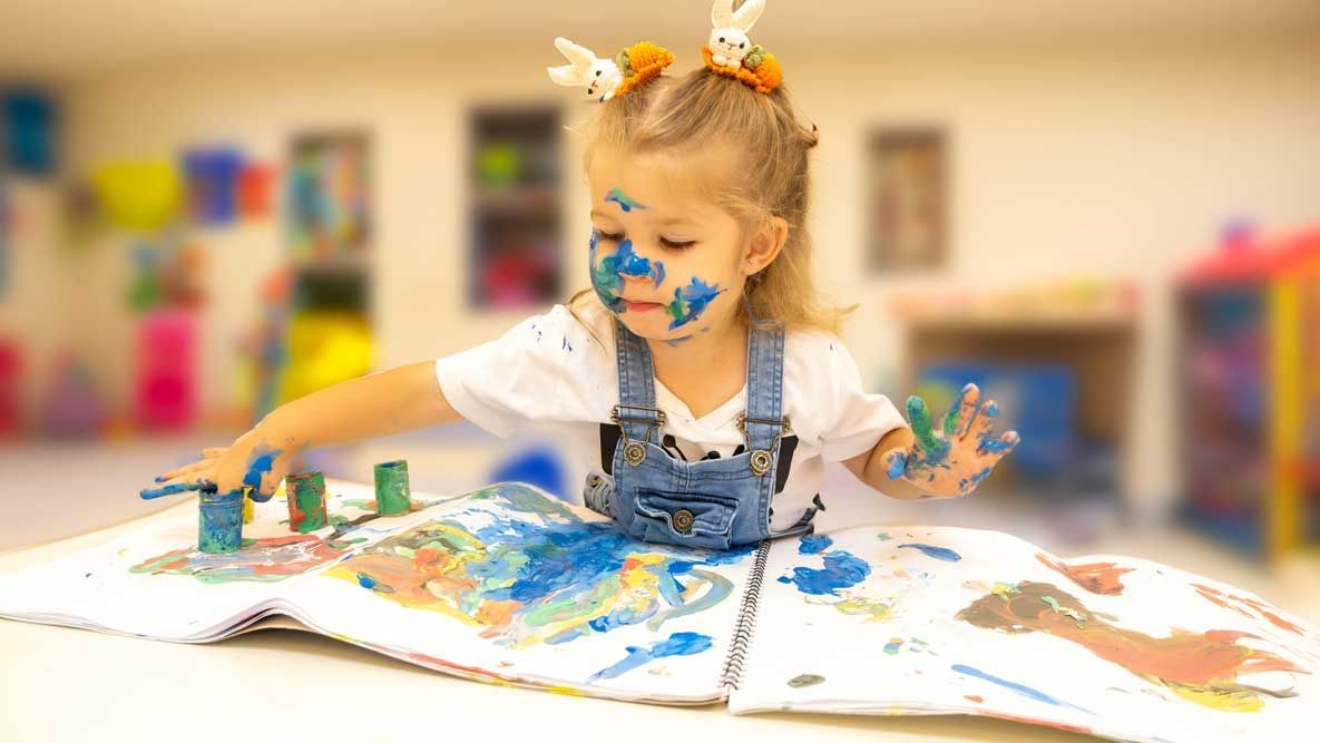 Child painting with colorful handprints and splashes at a table in a classroom