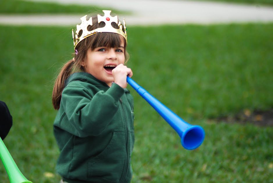 Child in a green hoodie blowing a blue party horn outdoors, wearing a paper crown.