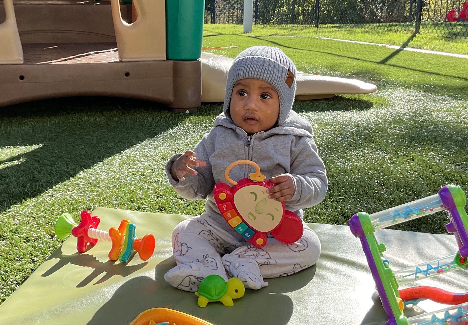 Baby sitting on a play mat outdoors with toys, wearing a gray knit hat and jacket.
