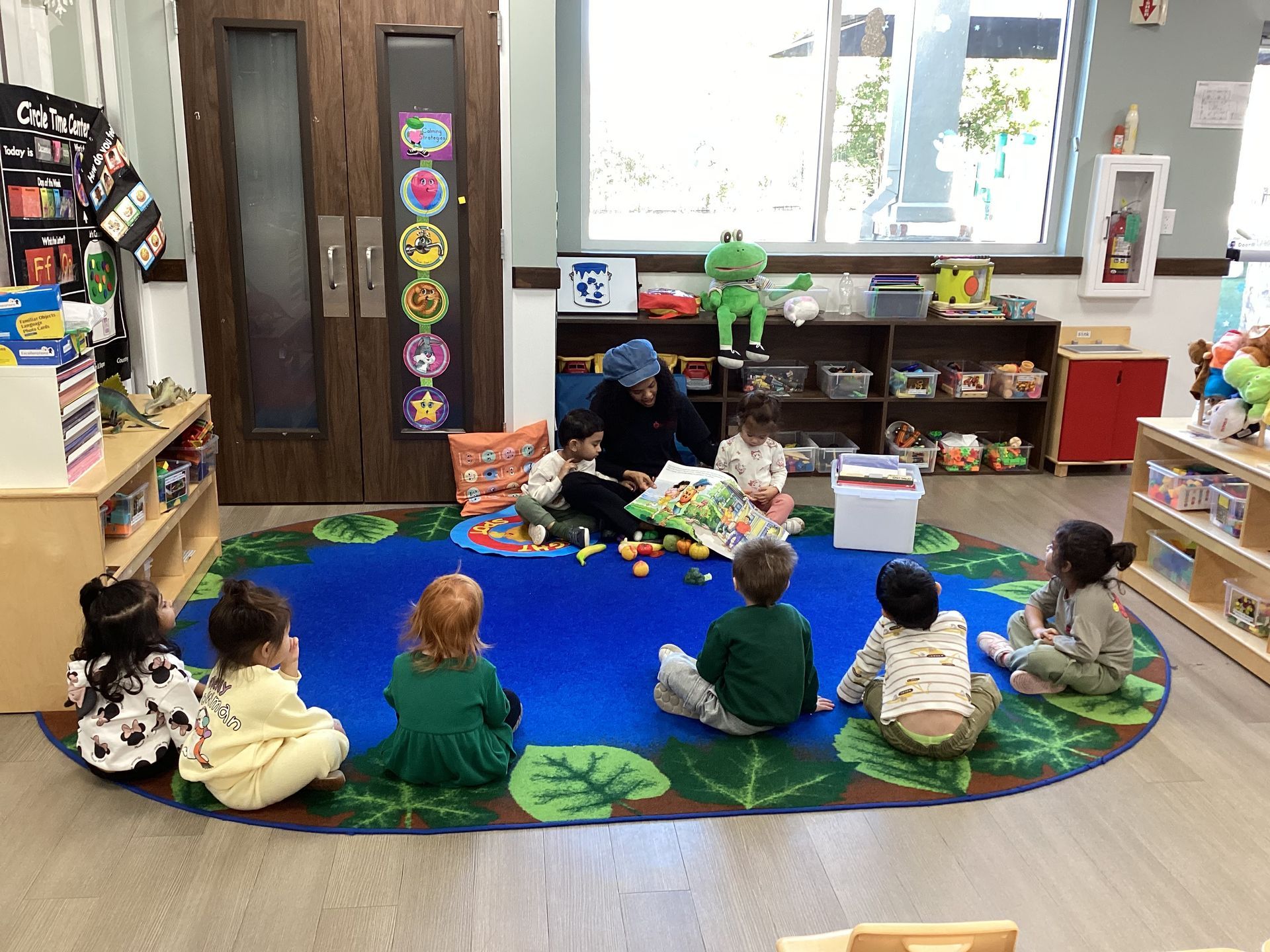 Children sit on a blue rug in a classroom, facing a teacher near shelves and toys.