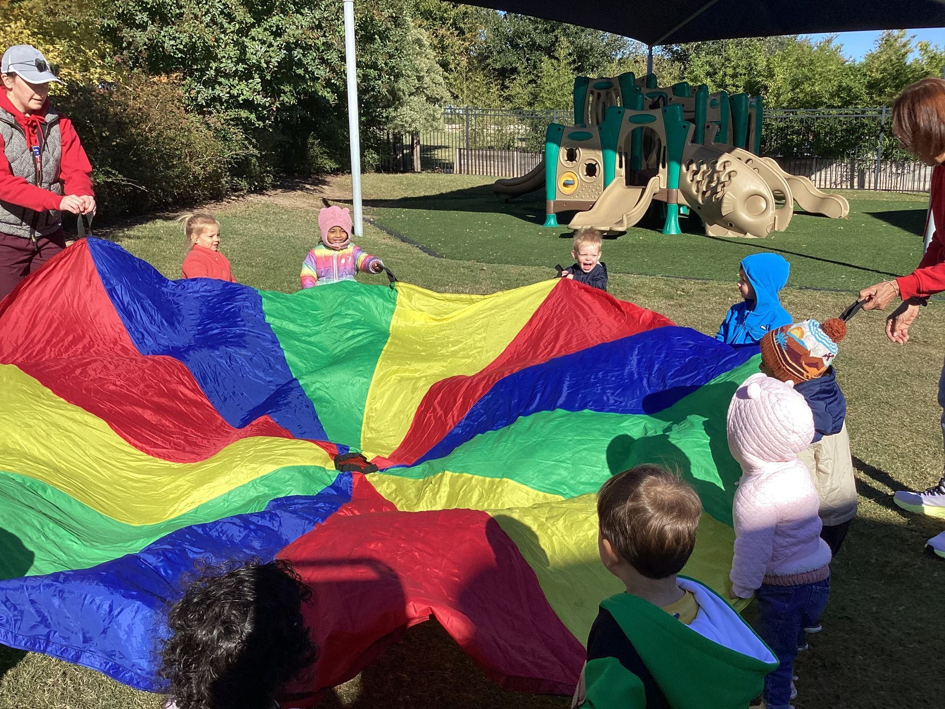 Children playing parachute game on a playground, holding a colorful red, blue, yellow, and green sheet