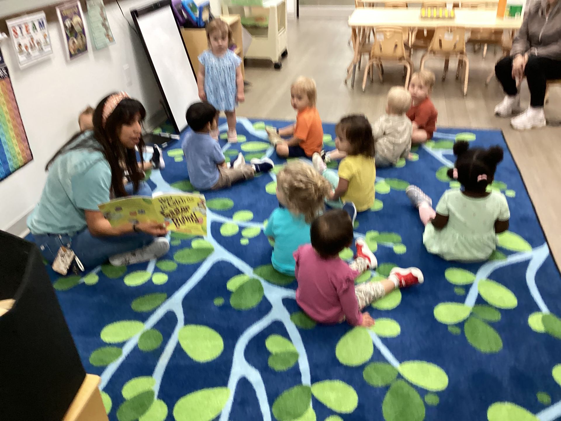 Children sitting on a blue-and-green leaf rug in a classroom, with bookshelves and toys around them.