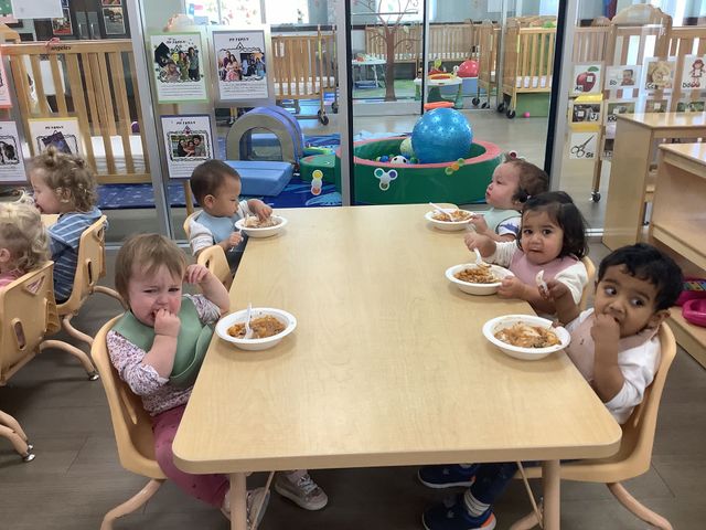 Children eating lunch at a table in a daycare classroom, with toys and play areas in the background.