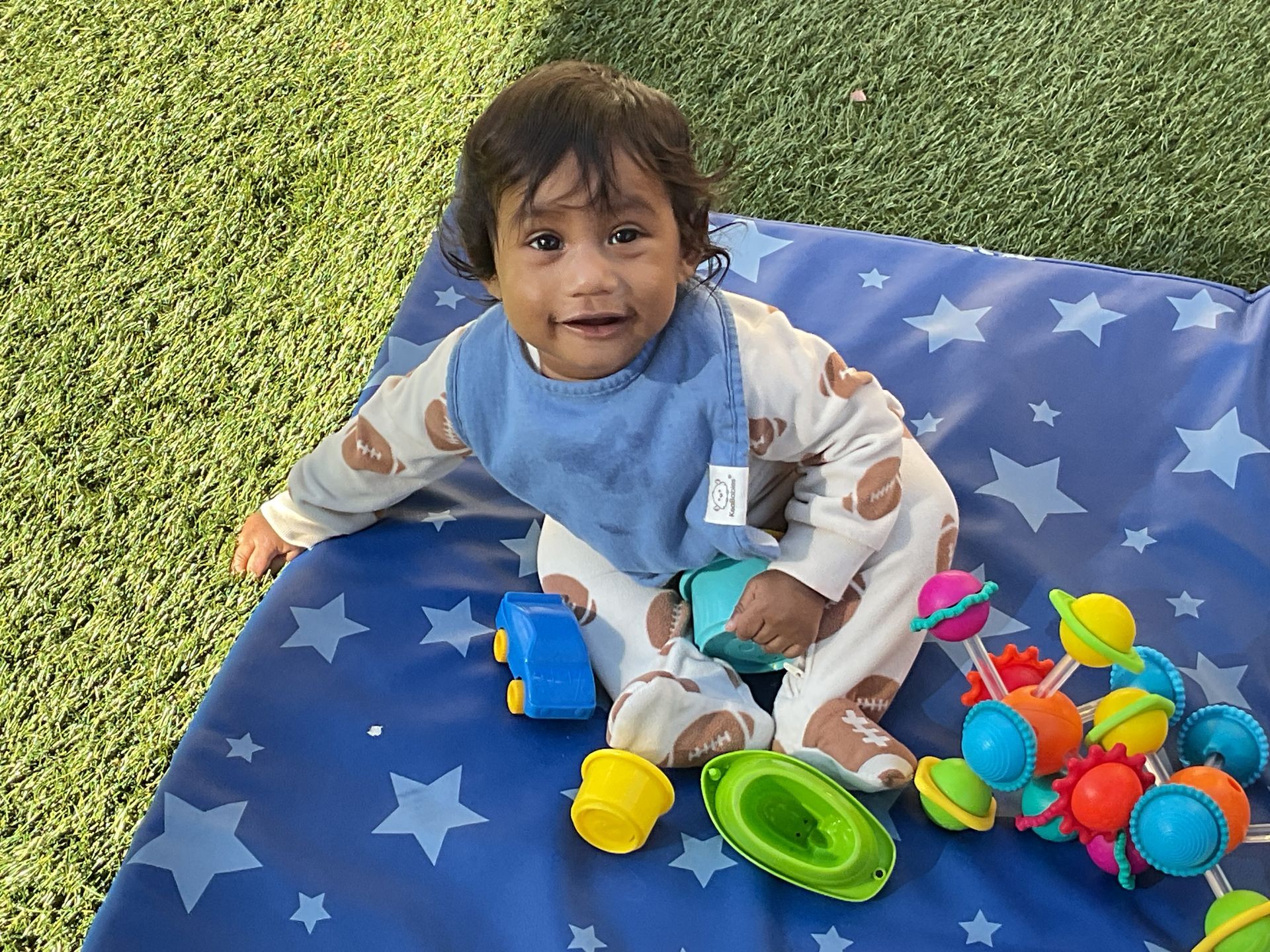 Child sitting on a blue star-pattern blanket with colorful toys on grass, smiling at the camera