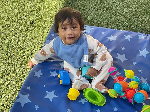 Toddler sitting on a blue star-pattern blanket with colorful toys on grass, smiling at the camera
