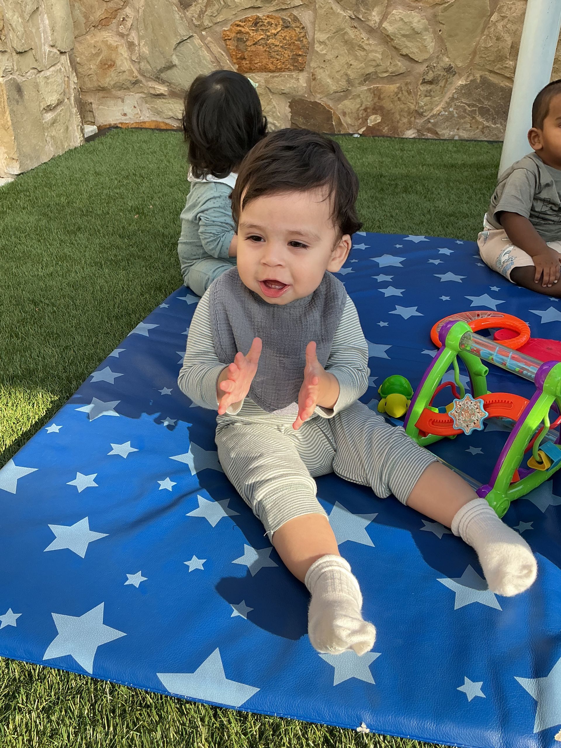 Toddler sitting on a blue star blanket, clapping near colorful toys outdoors.