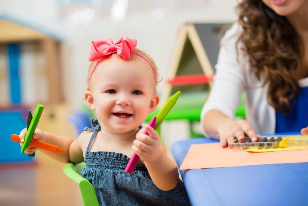 Smiling toddler holding colorful markers at a craft table, with an adult helping in the background