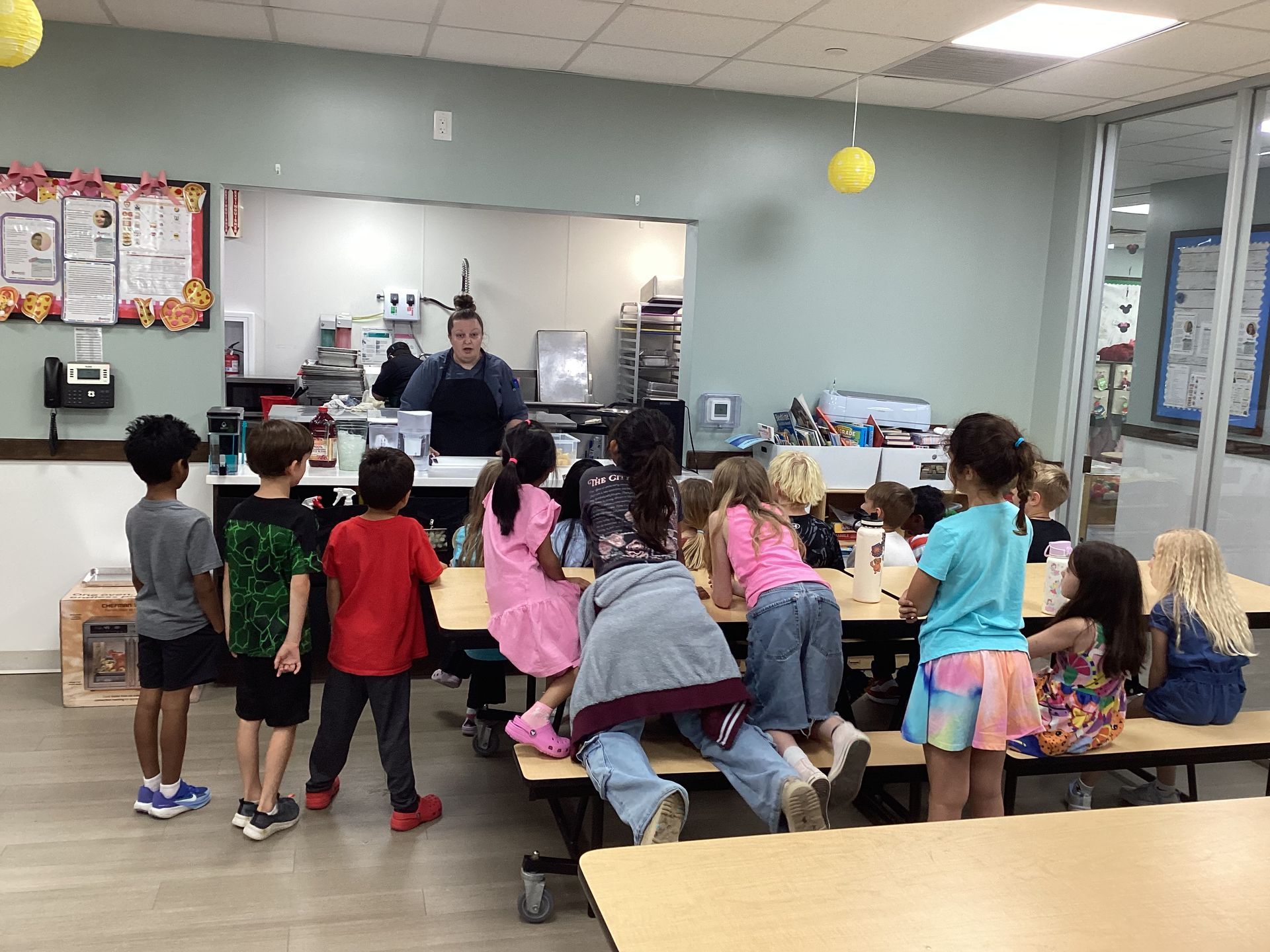 Children gathered around tables in a classroom while a teacher stands at the front.