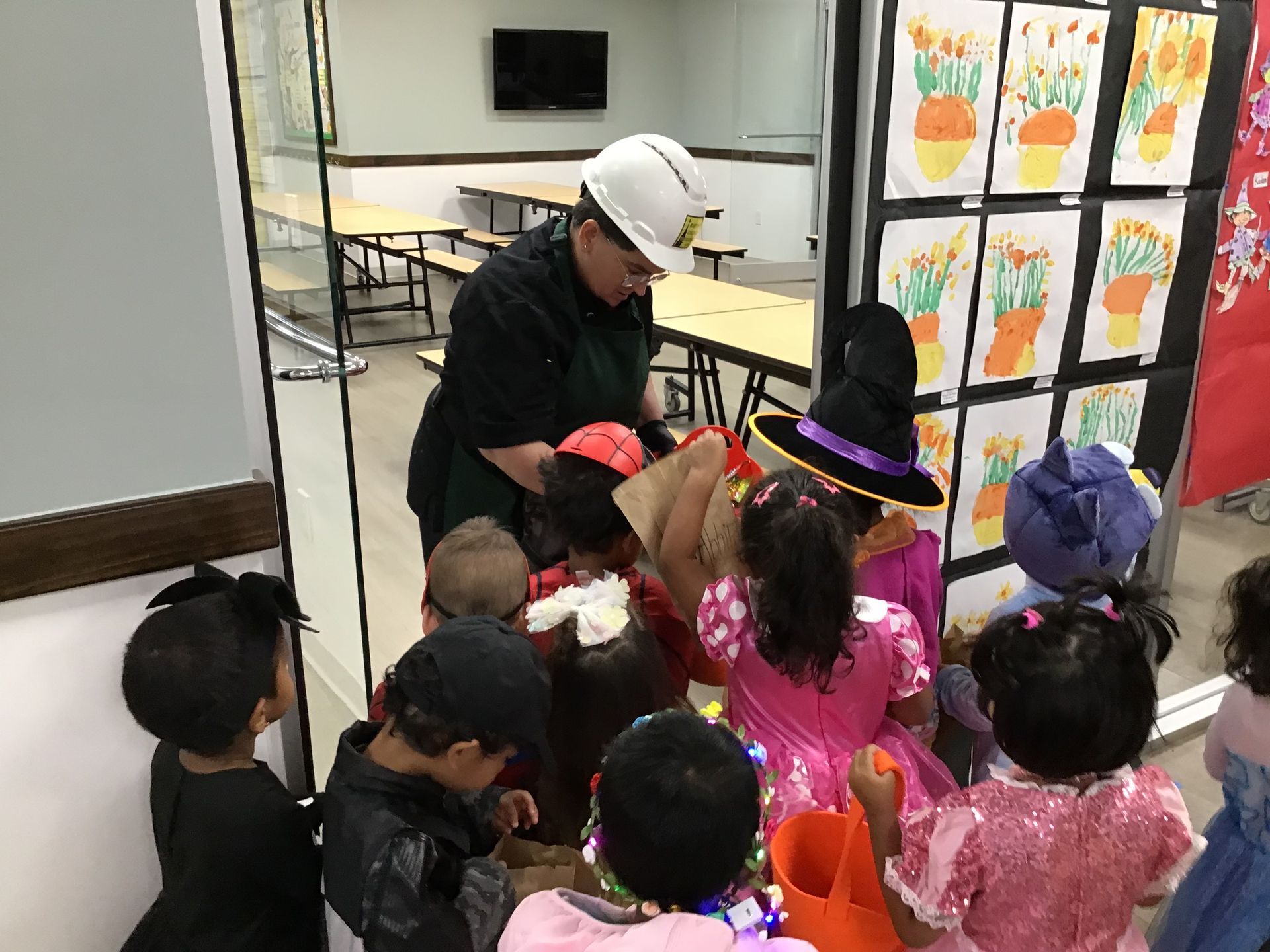 Children in costumes gather around an adult in a classroom, facing a wall of colorful art drawings.