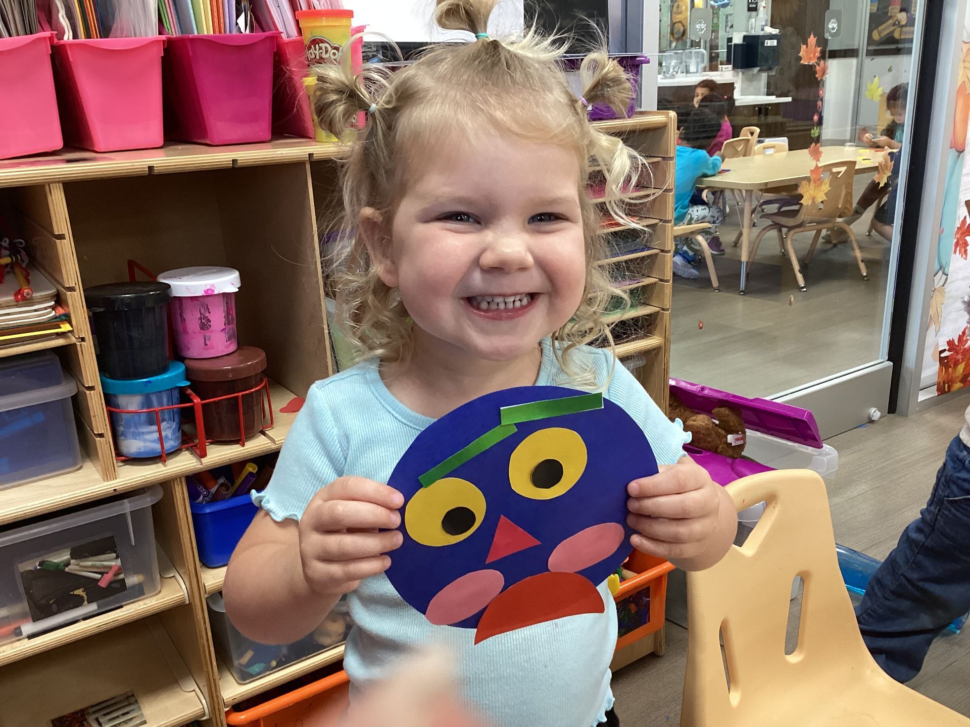 Smiling child holding a colorful owl craft in a classroom craft area