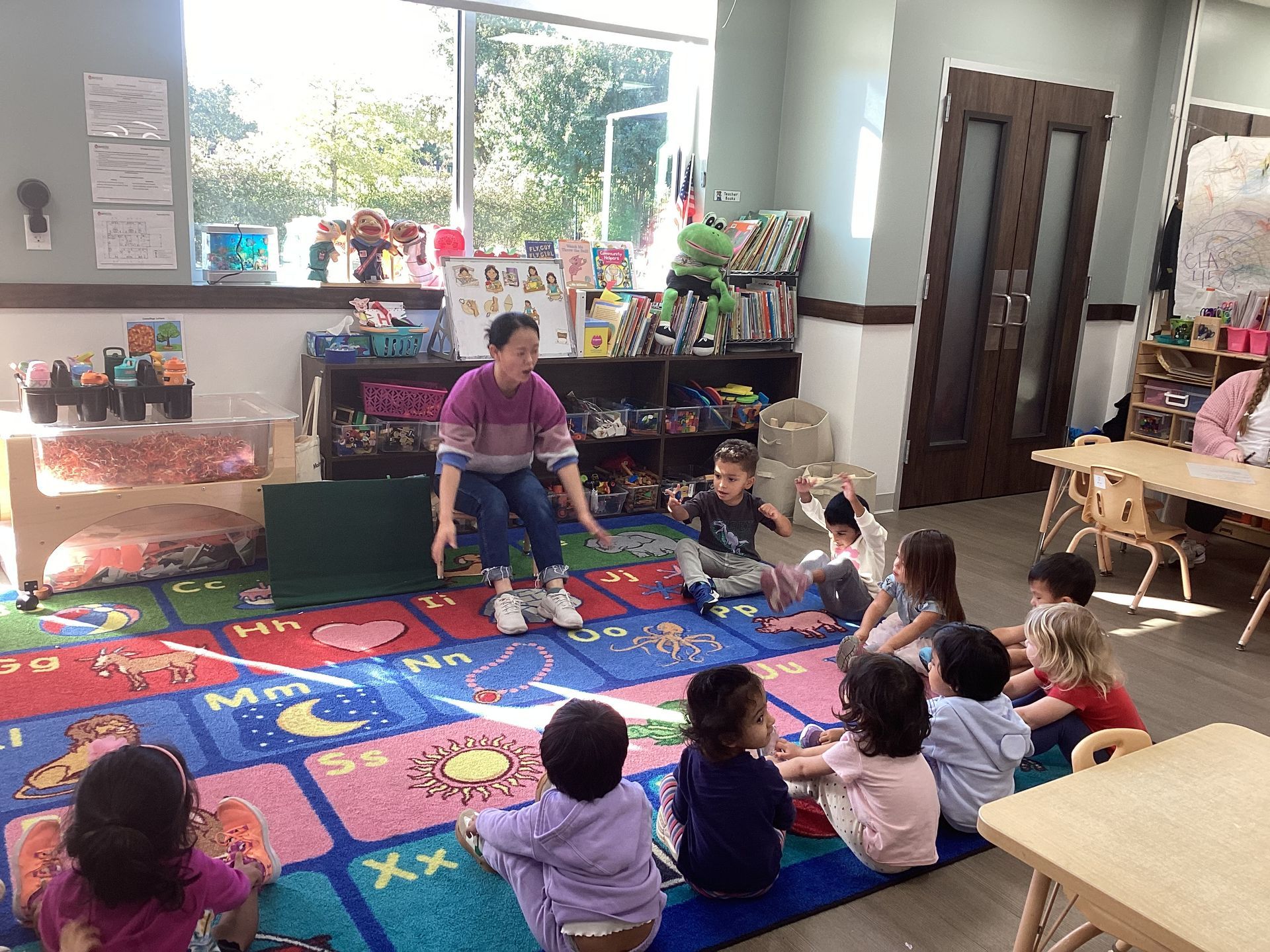 Preschool children sitting on a colorful rug with a teacher in a classroom near a bright window