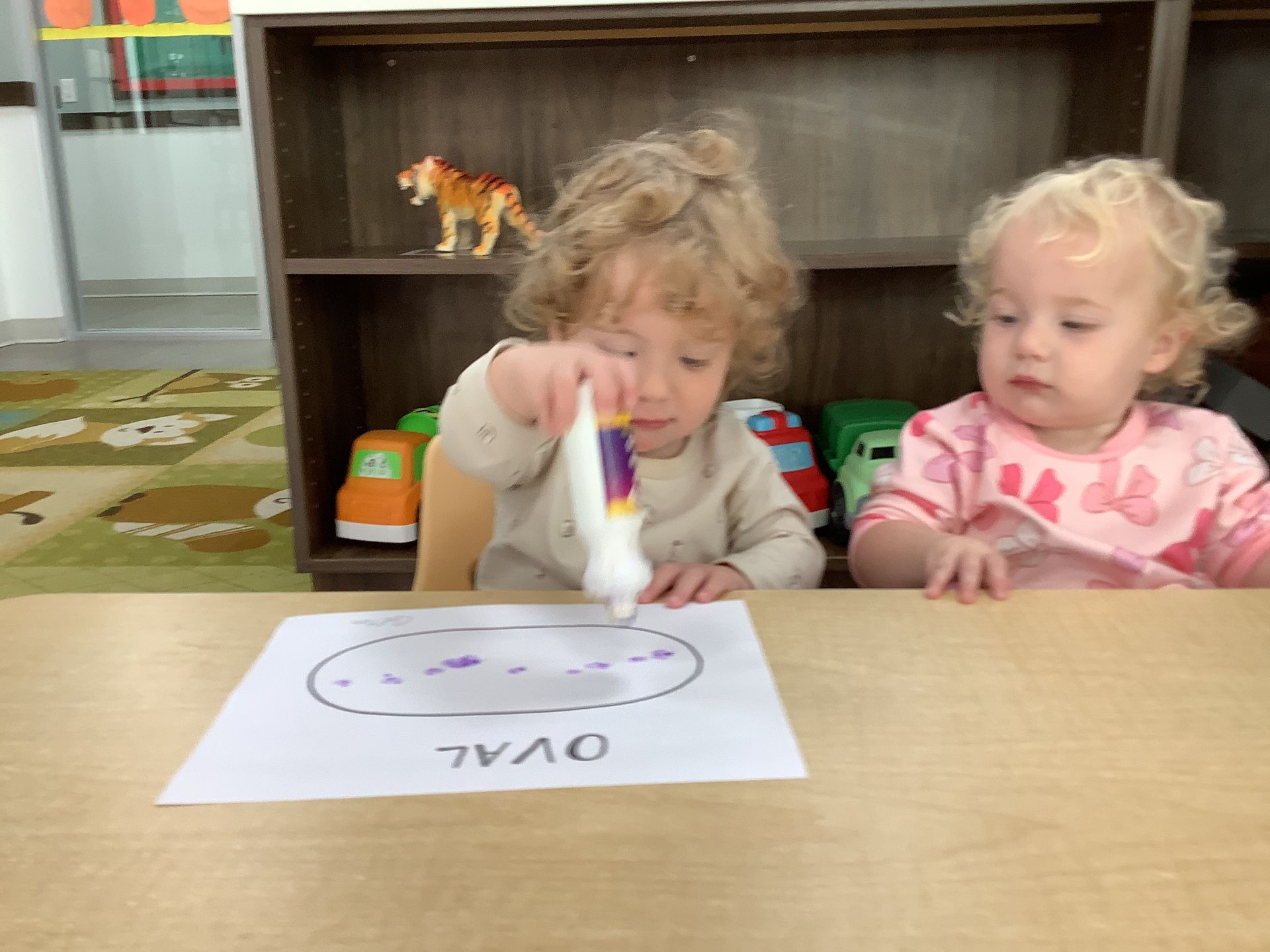 Two toddlers at a table, drawing on paper with markers in a classroom.