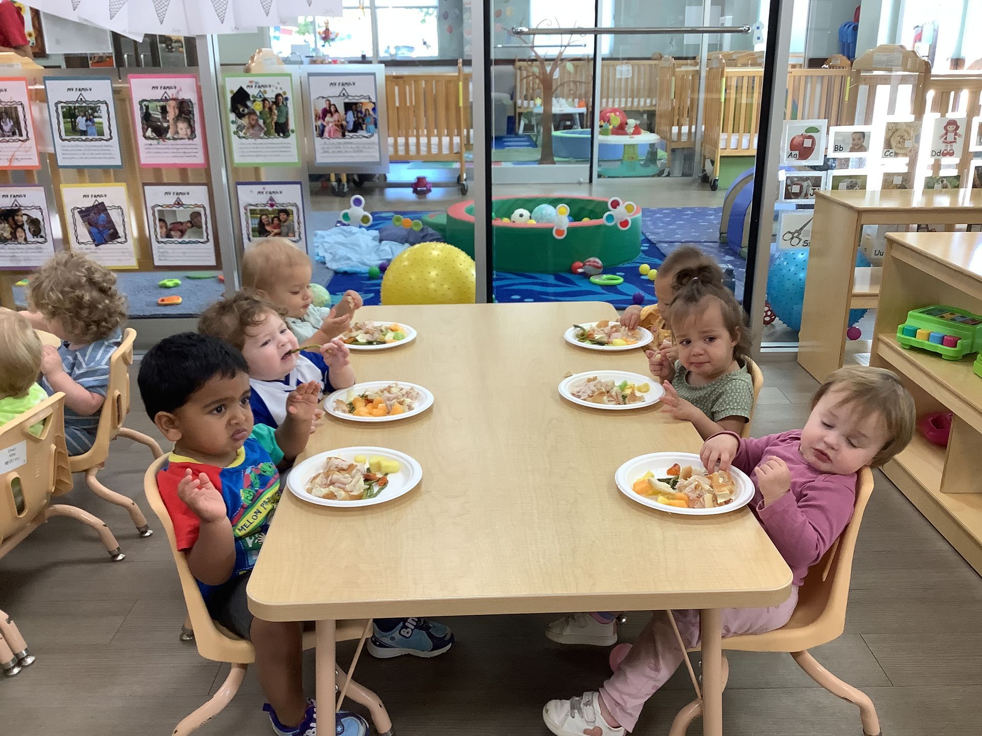 Children eating at a daycare table in a bright playroom, with plates of food and toys in the background