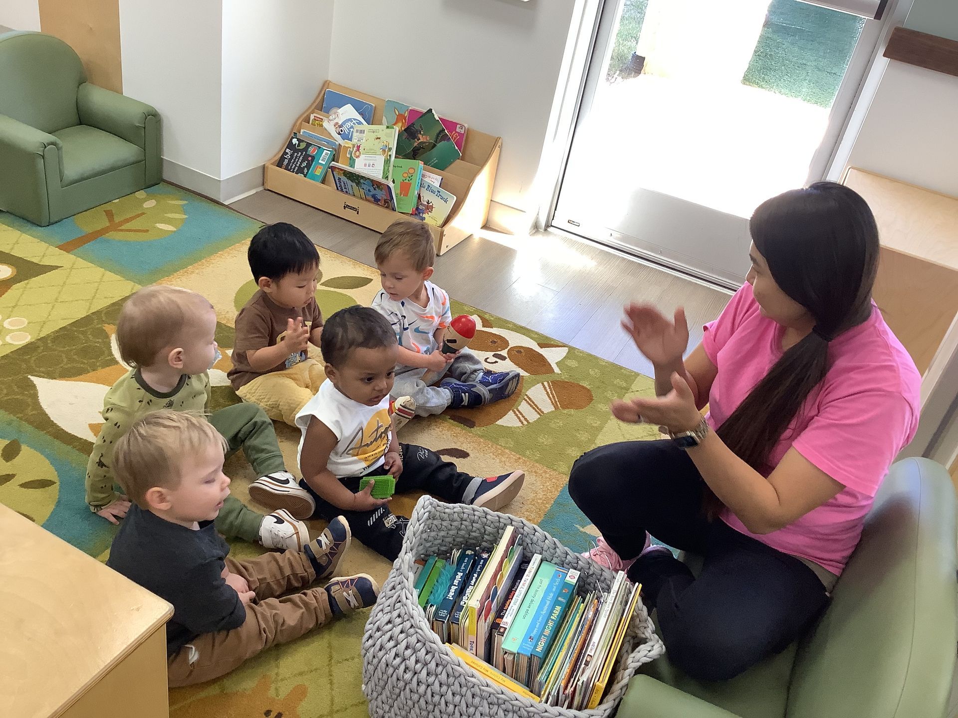 Caregiver in pink sits with a small group of children on a classroom rug, clapping during circle time.