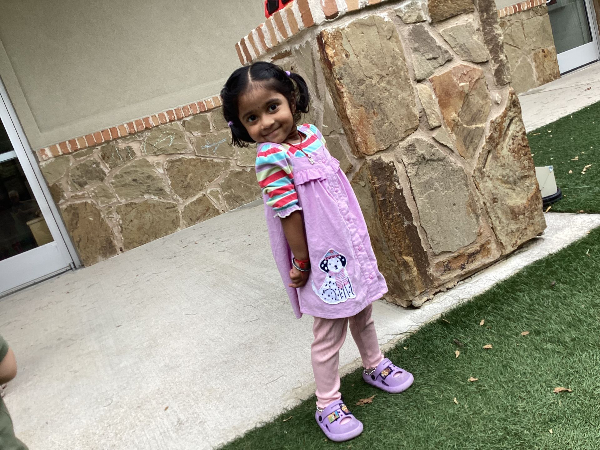 Child in a pink outfit standing on a sidewalk near a stone column, smiling at the camera.