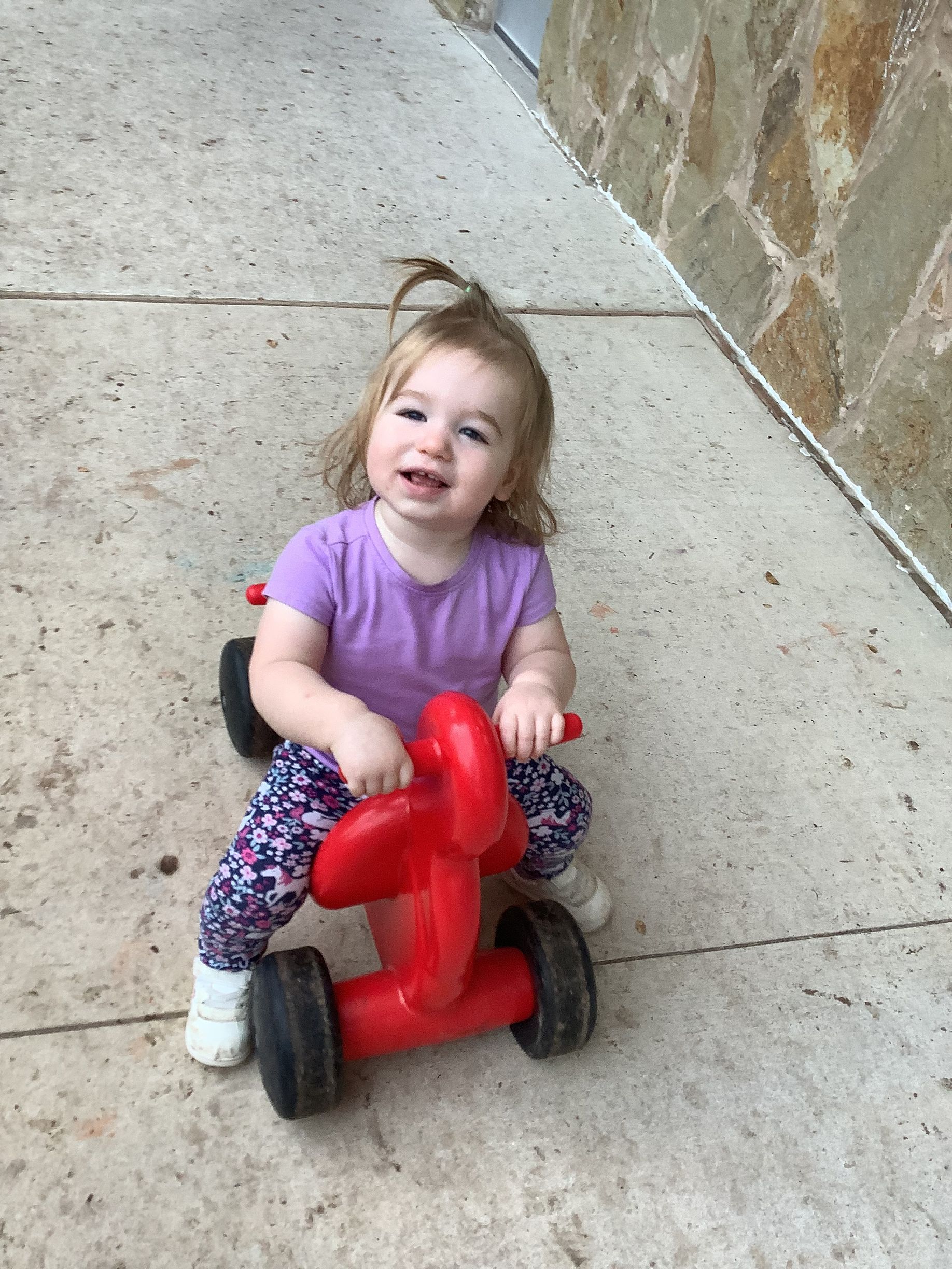 Smiling child on a red ride-on toy on a concrete sidewalk beside a stone wall