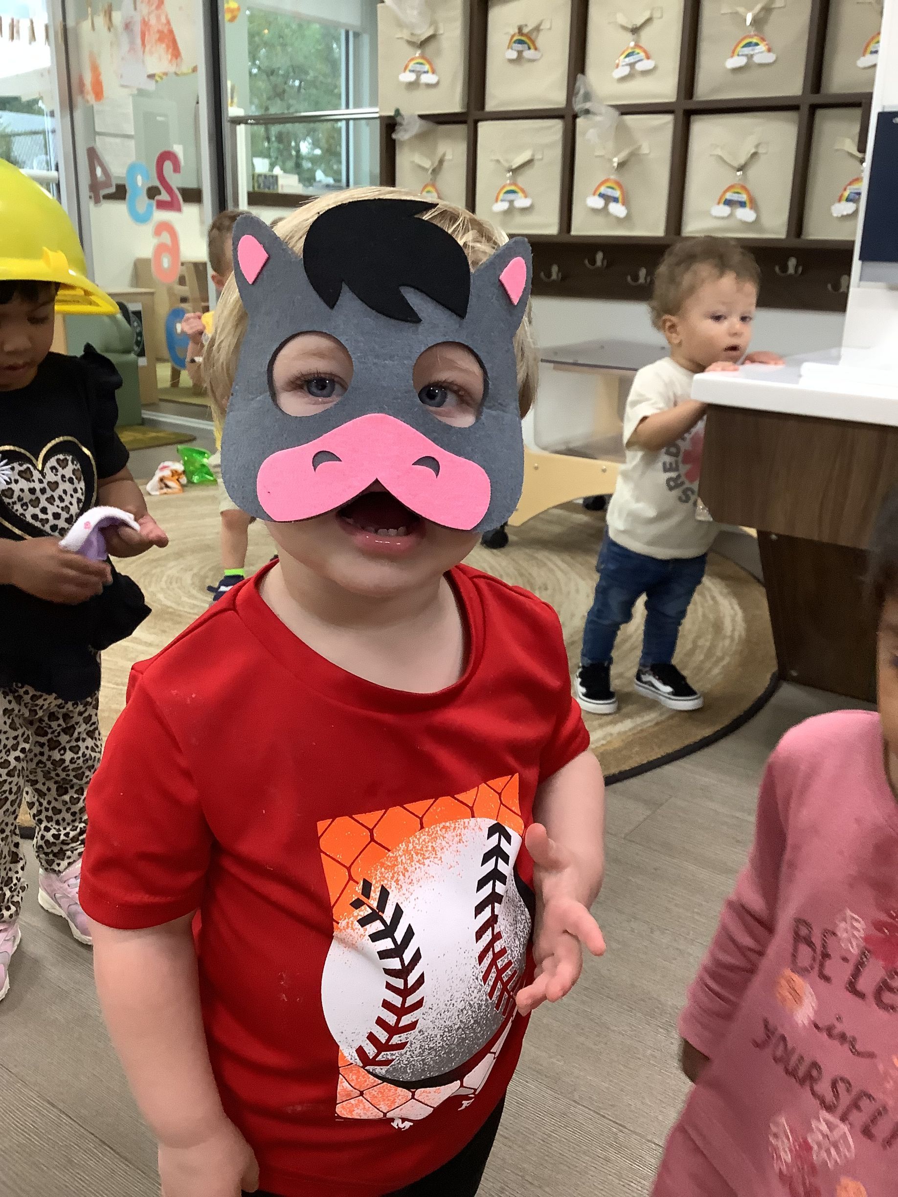 Child wearing a black cat mask and red shirt in a classroom, with other children in the background