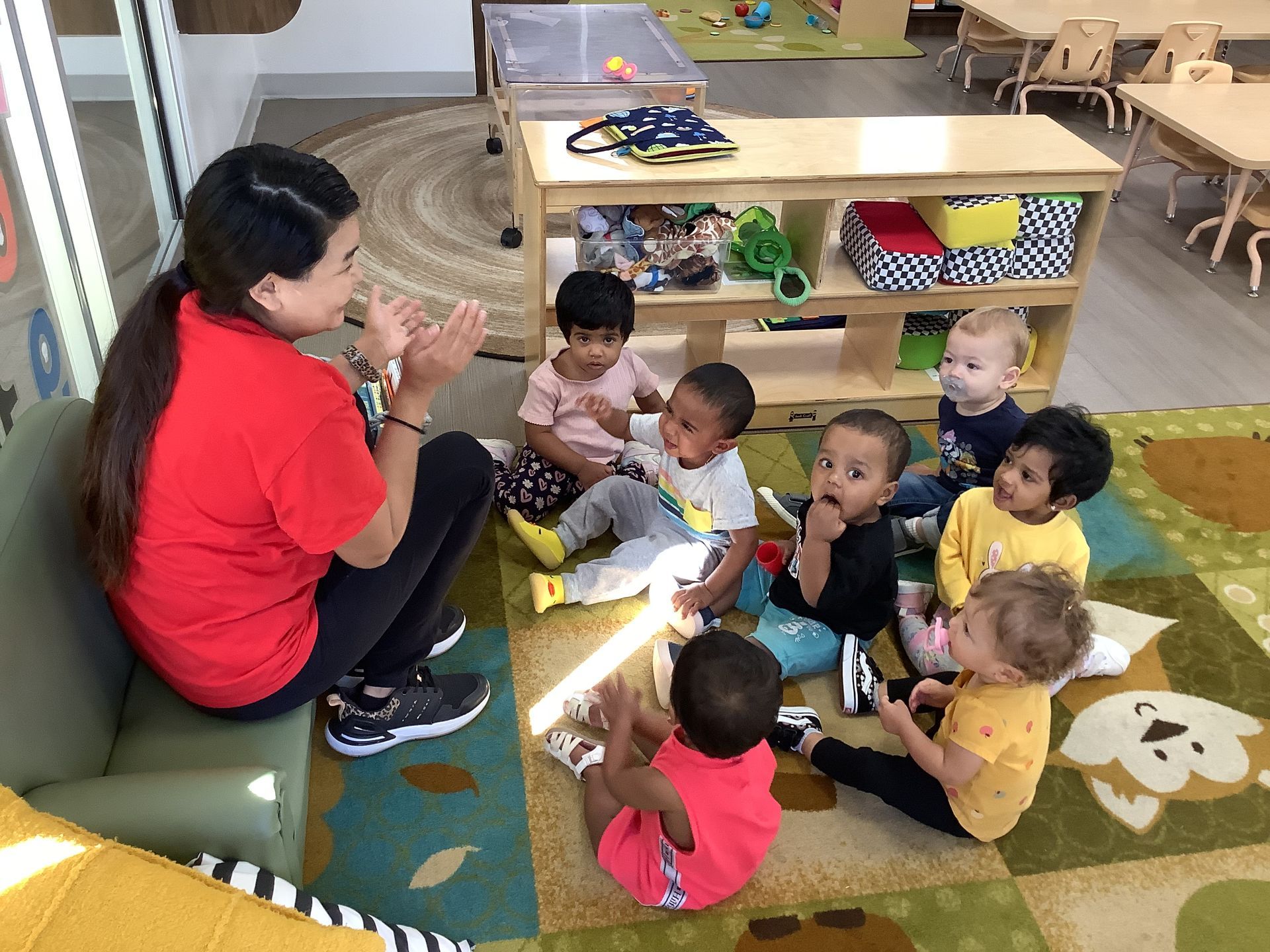 Teacher in red shirt sits with toddlers on a colorful classroom rug, leading a small group activity.