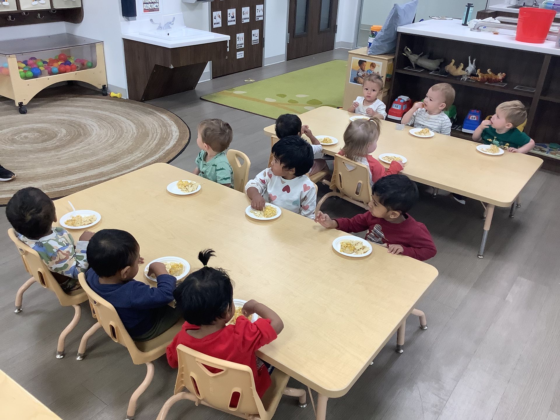 Children seated around tables in a daycare classroom, eating snacks and drinks.