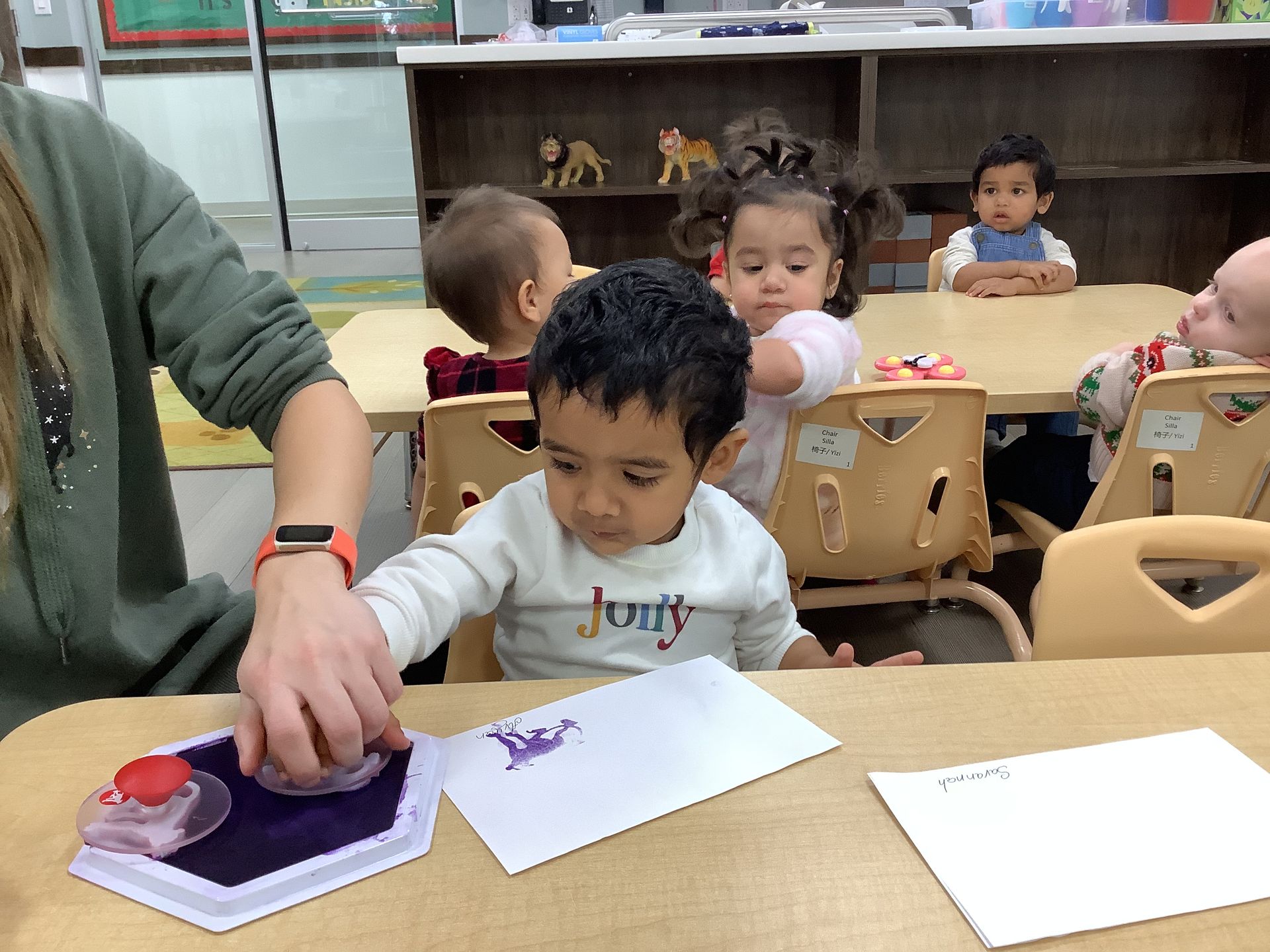 Child painting at a classroom table while an adult helps, with other children seated in the background.