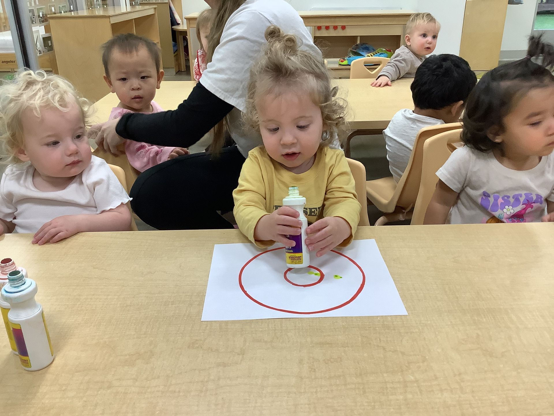Children in a classroom painting at a table, with one child drawing a red circle on paper