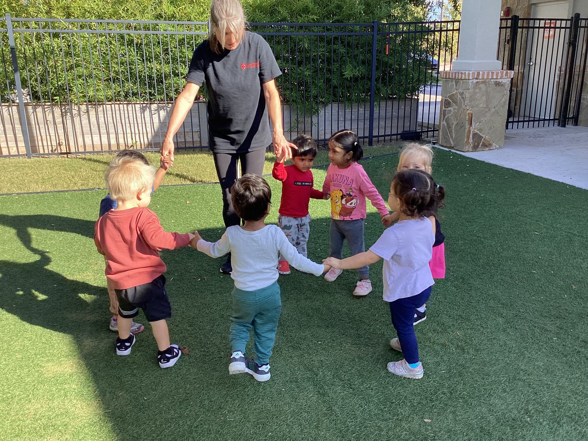 Teacher leading toddlers holding hands in a circle on a playground turf area