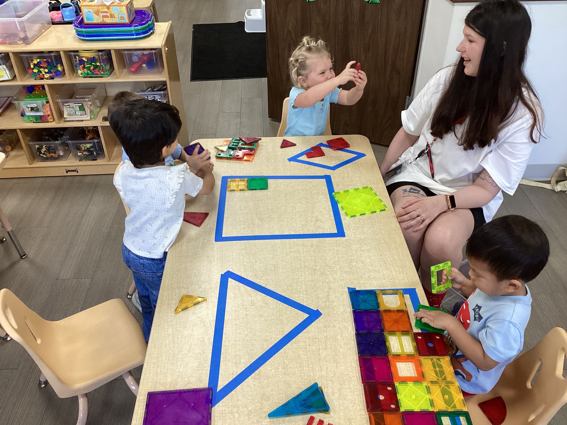 Children and a teacher sit around a table with colorful shape puzzles in a classroom.