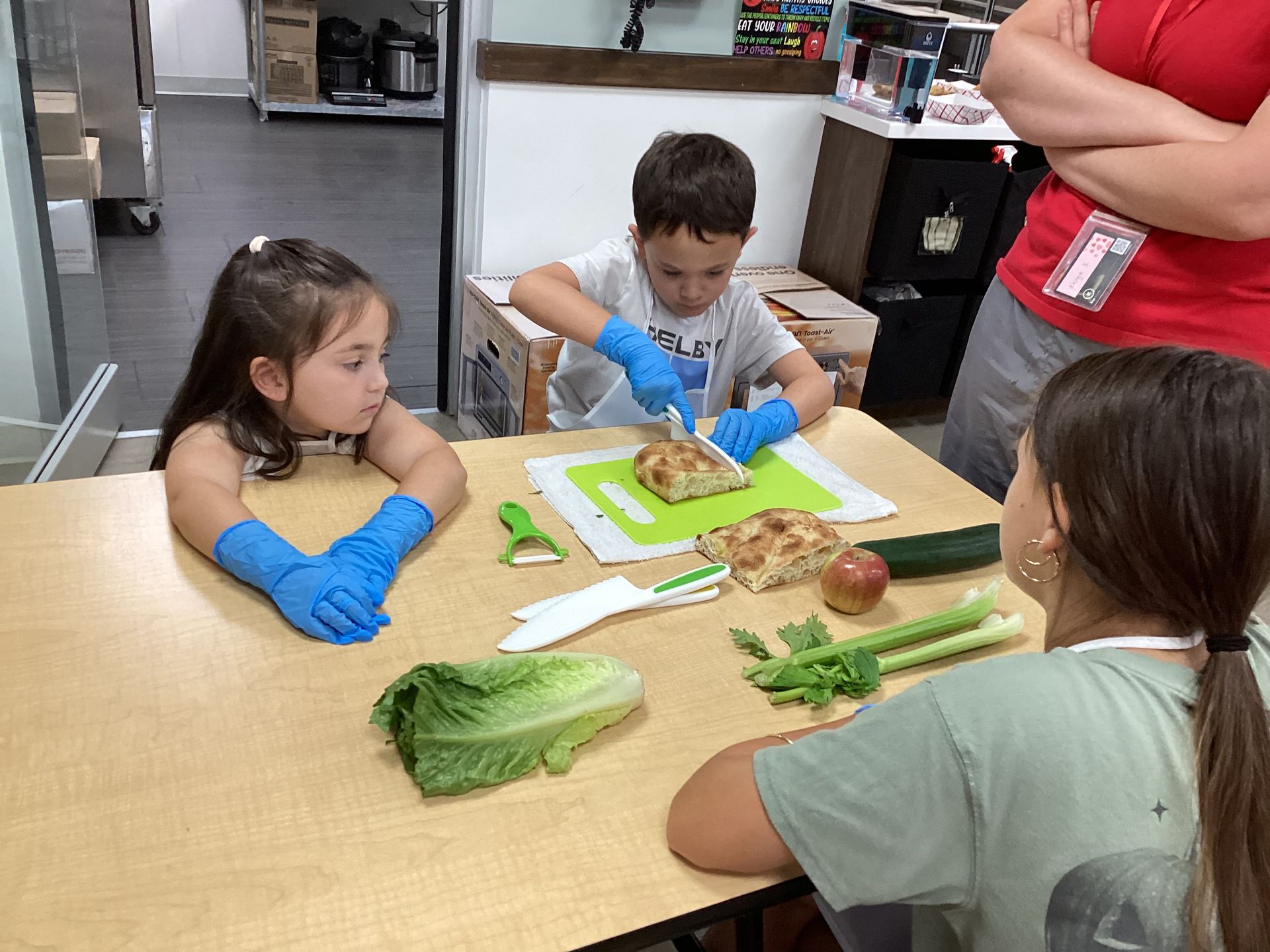 Children wearing blue gloves prepare vegetables at a kitchen table during a cooking activity